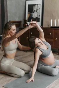 A blonde woman assists a pregnant woman in gray athletic wear with a side stretch on a yoga mat.