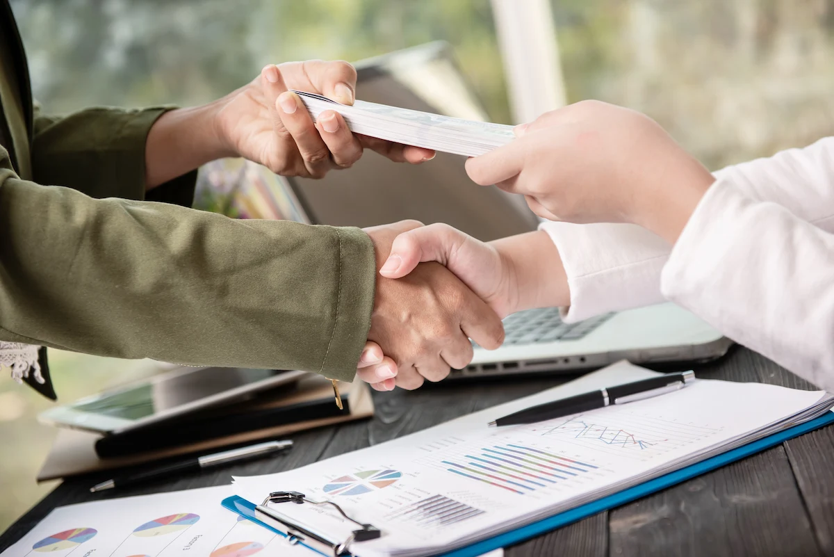 Two people shaking hands and exchanging money over a desk with business charts and a laptop.