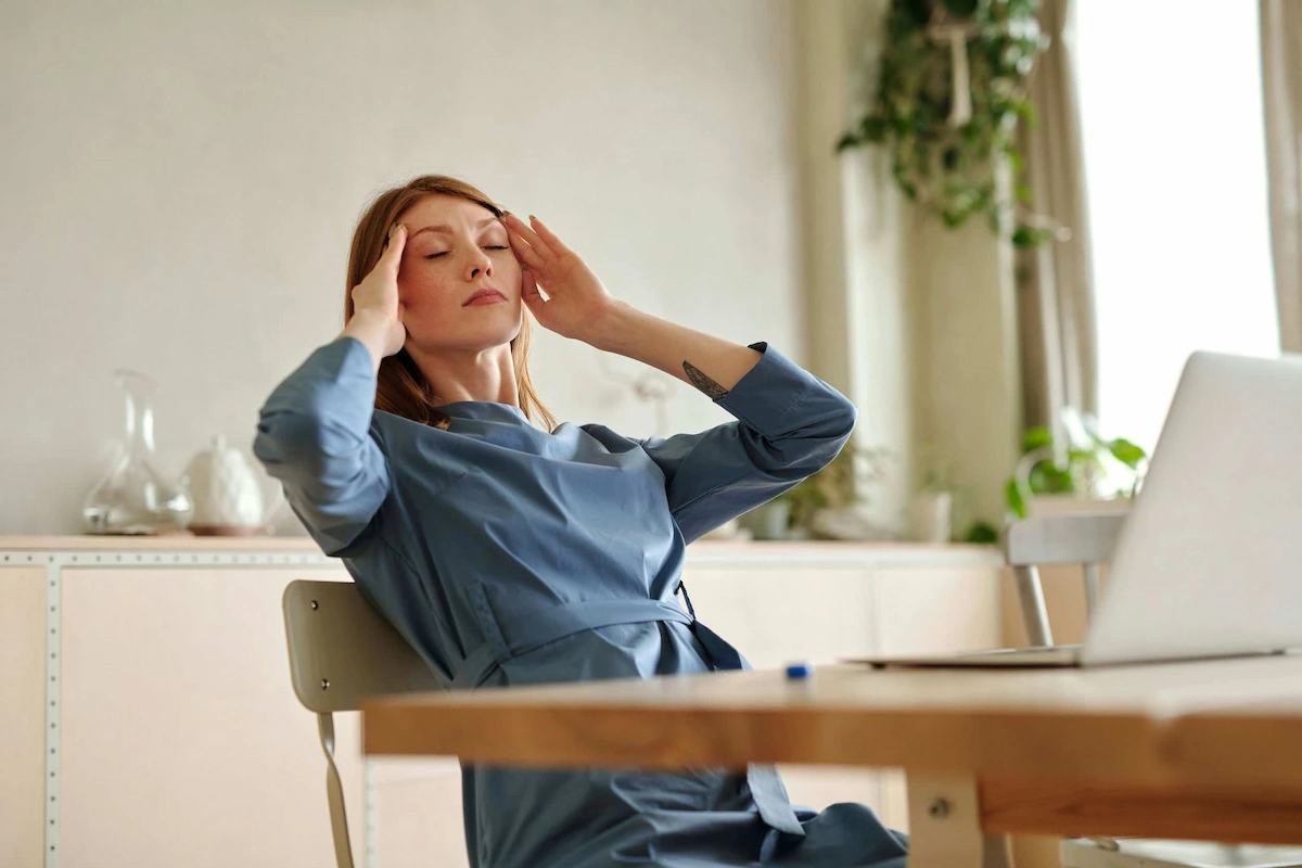 A woman in a blue dress with closed eyes presses her temples, appearing to have a headache, at a desk with a laptop.