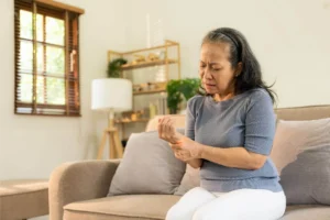 An older woman with a pained expression sits on a couch, holding her left wrist with her right hand.