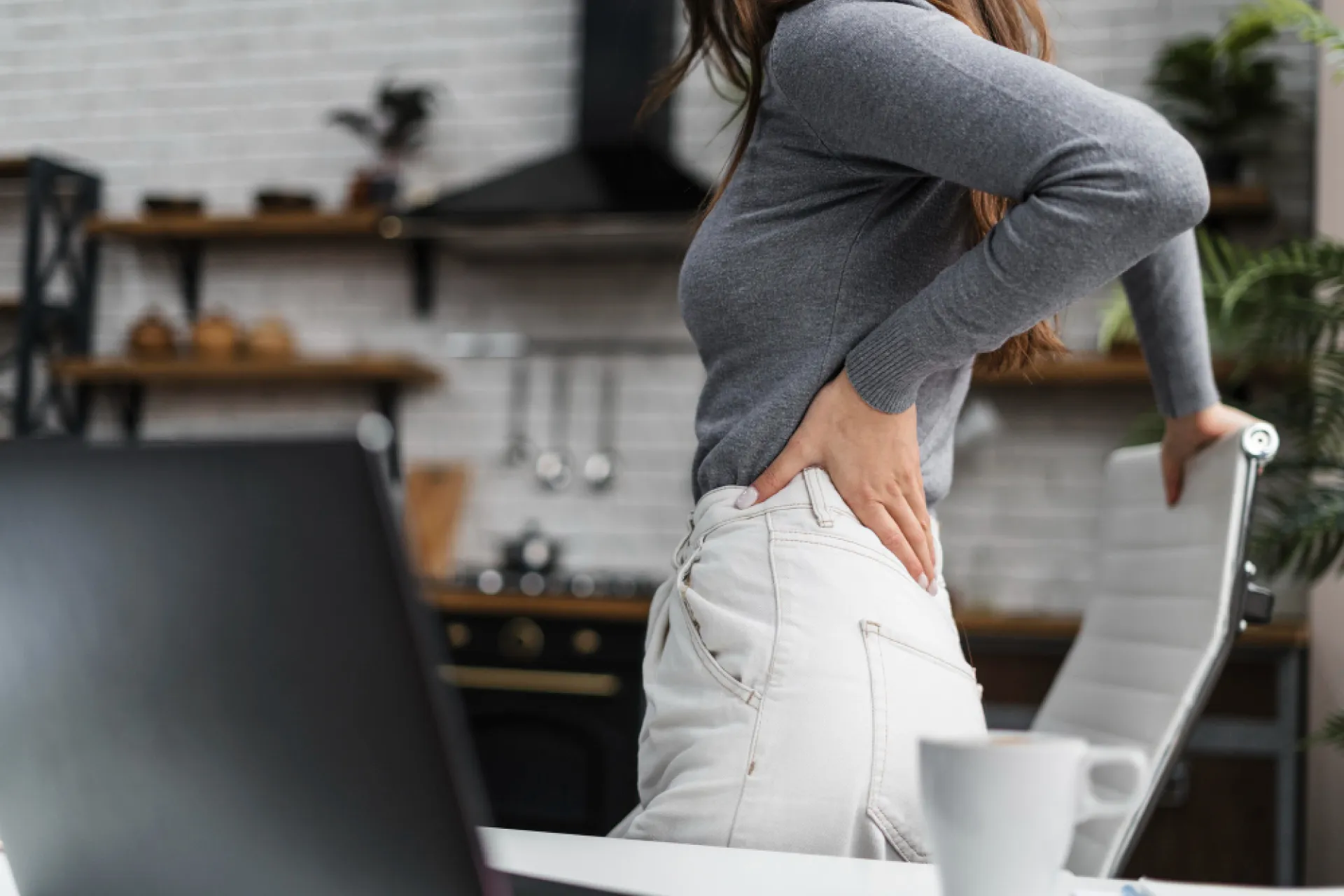 A woman in a grey top and light jeans stands, holding her lower back in discomfort, next to a white office chair and a laptop in a home kitchen setting.