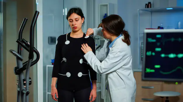 A medical professional in a lab coat attaches electrodes to a patient wearing a black top, with an elliptical machine and medical monitor in the background.