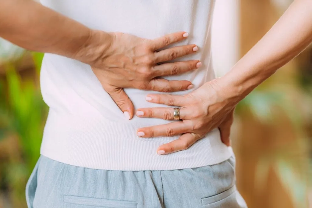 A person from behind, wearing a white top and light pants, holding their lower back with both hands, suggesting back pain.