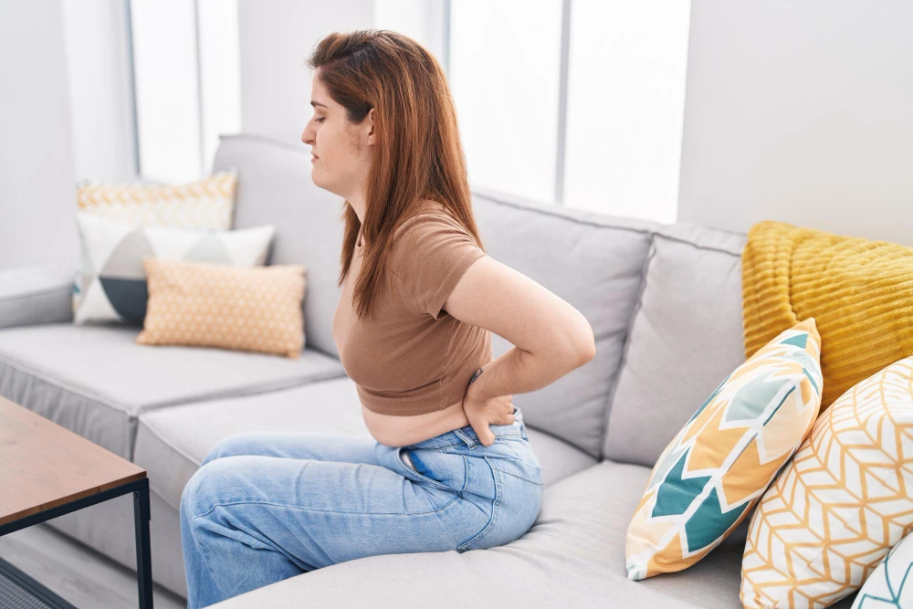 A woman sits on a gray sofa, holding her lower back with both hands, eyes closed in discomfort.