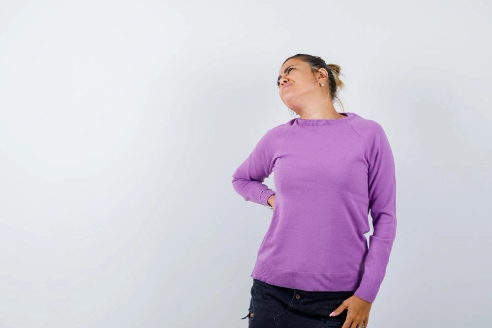 A woman in a purple sweater and dark skirt, looking up with a strained expression, holding her lower back with one hand.