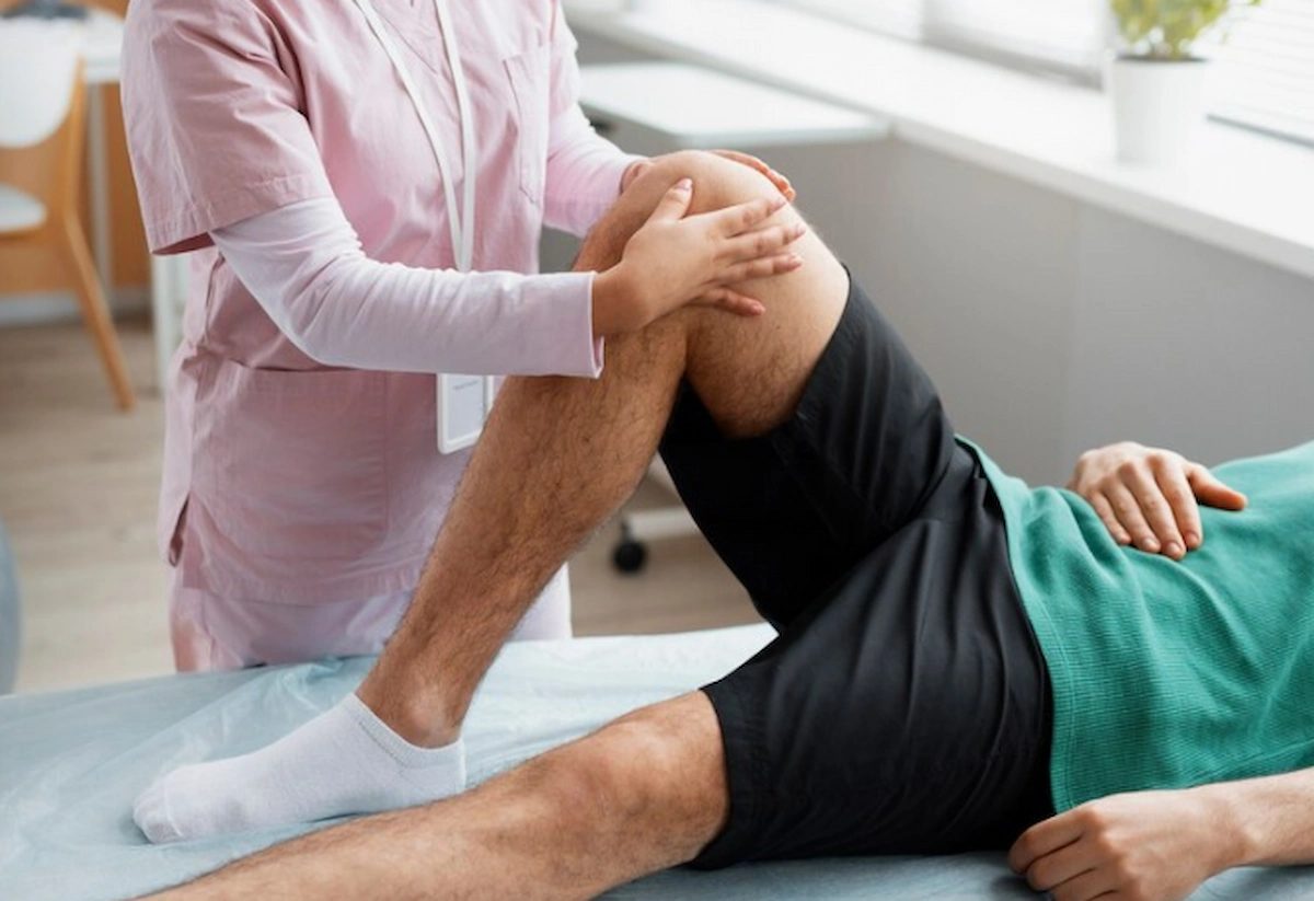 A medical professional in pink scrubs examines a patient's bent knee on an examination table.