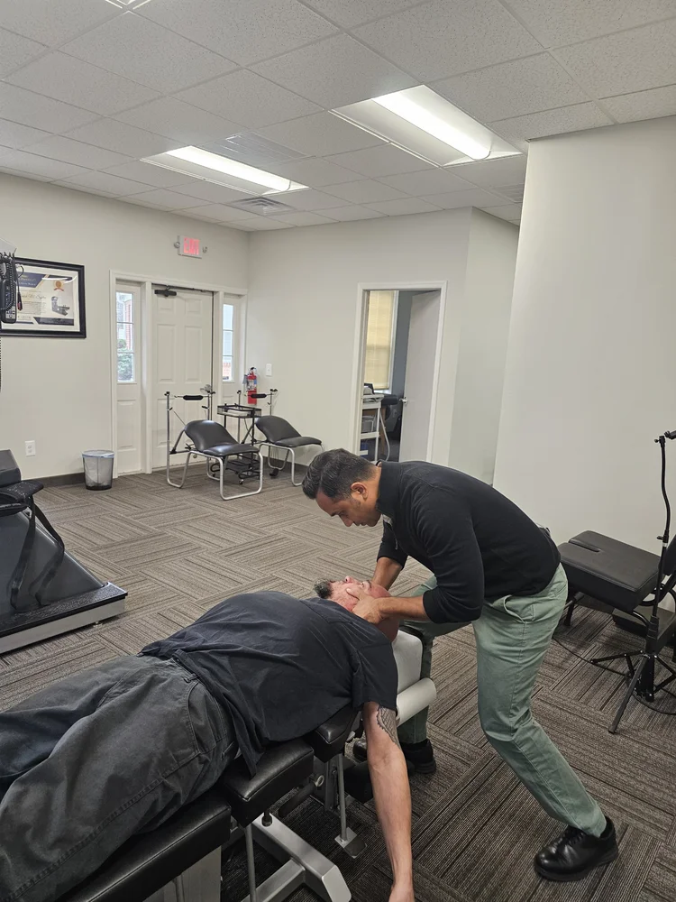A chiropractor in a black shirt and green pants adjusts the neck of a patient lying face up on a black examination table in a medical office.