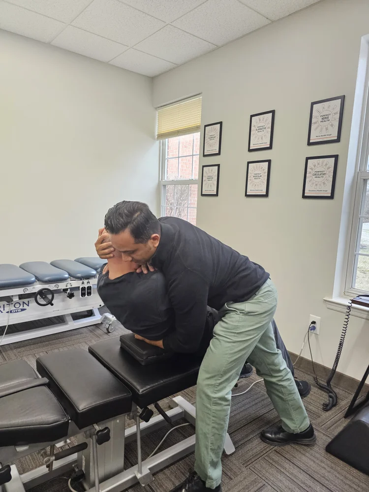 A chiropractor adjusts a patient's upper back/neck on a black chiropractic table in a clinic with white walls and framed wellness posters.