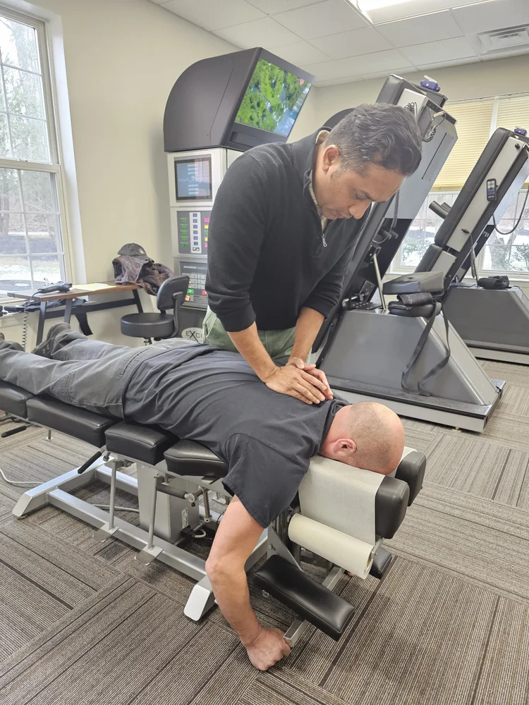 A chiropractor adjusts a patient's upper back on a chiropractic table in a clinic setting, with medical equipment and a window in the background.