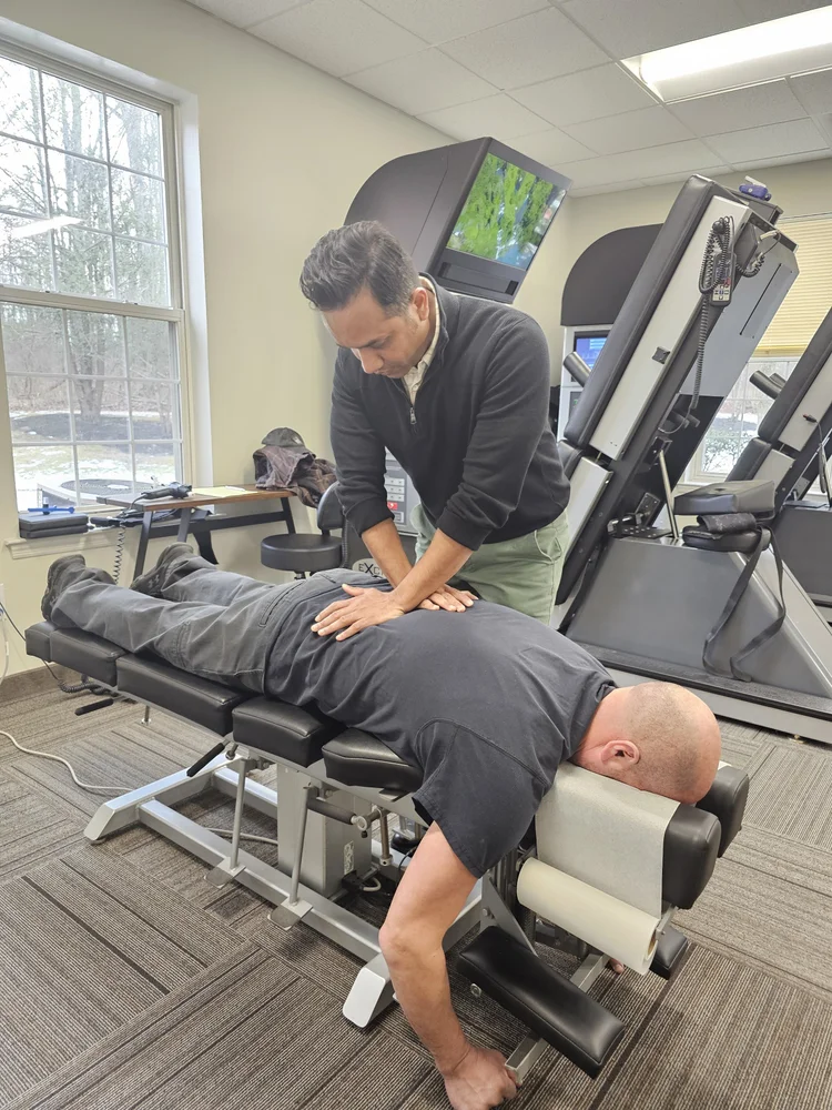 A chiropractor adjusts a male patient's lower back as the patient lies face down on a chiropractic table in a clinic.
