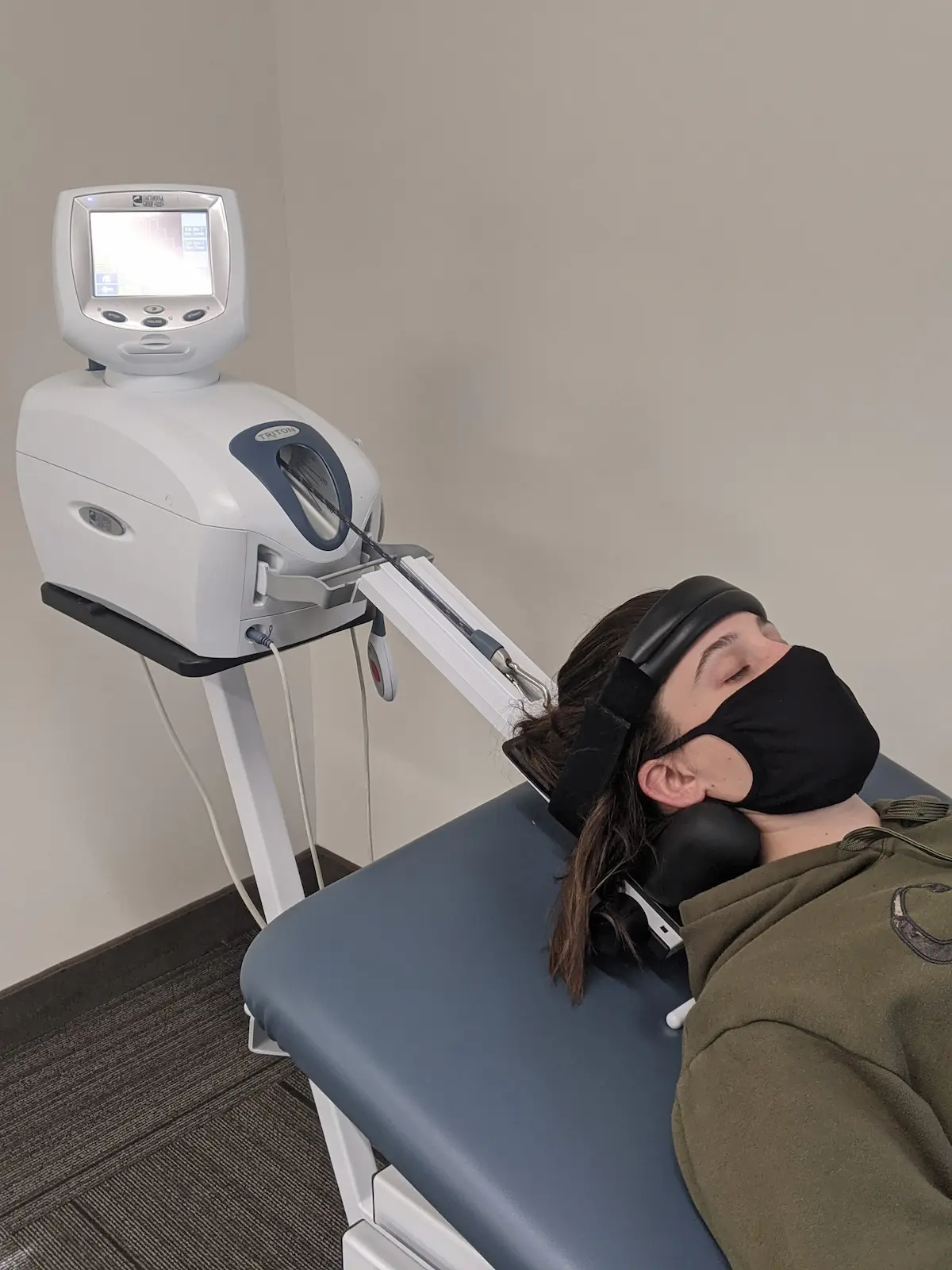 A person wearing a black face mask lies on a blue medical table, connected to a white Triton DTS spinal decompression machine via a head harness and mechanical arm.