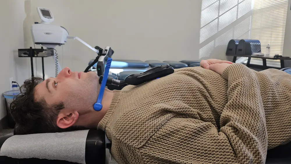 A man lies on a treatment table with a medical device attached to his head and neck, wearing a brown sweater.