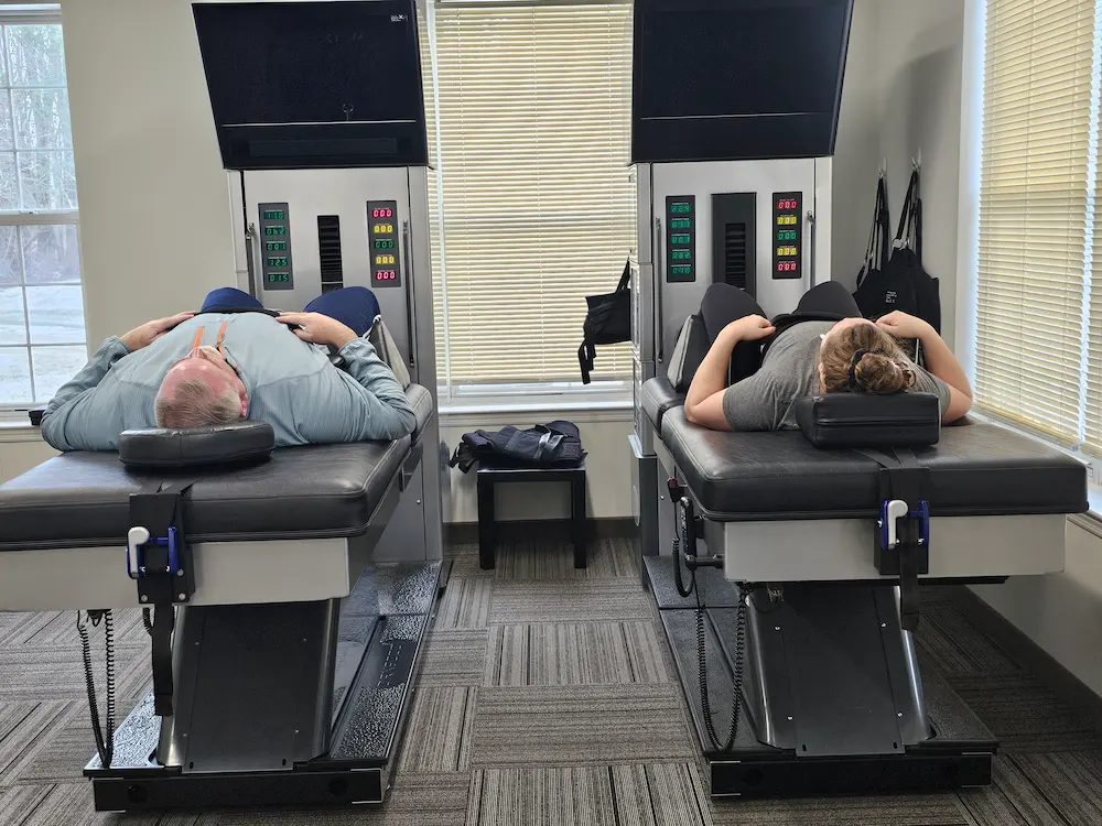 A man and a woman lie face up on separate spinal decompression tables in a therapy room.