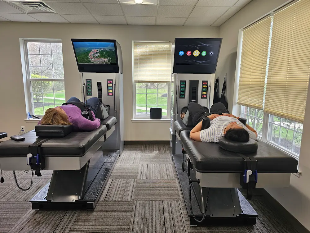 Two people lie face up on black and grey spinal decompression tables, each with a screen above showing different content, in a bright room with windows.