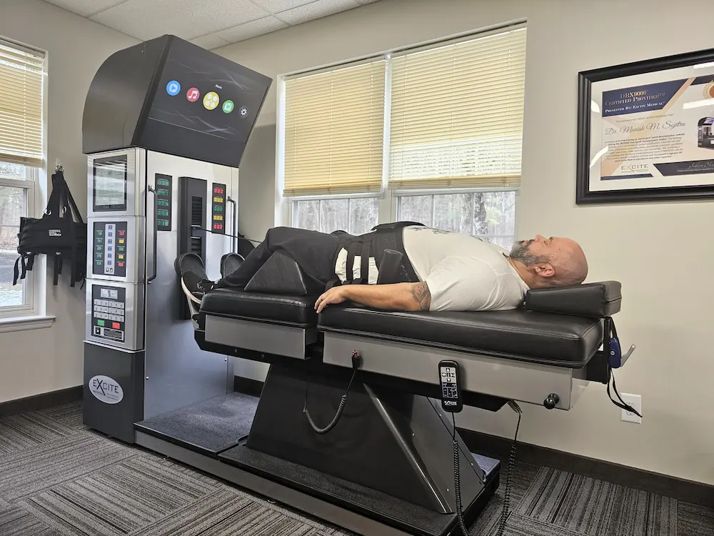 A man lies on his back on a large medical machine with straps, likely for spinal decompression, in a room with windows and a framed certificate.