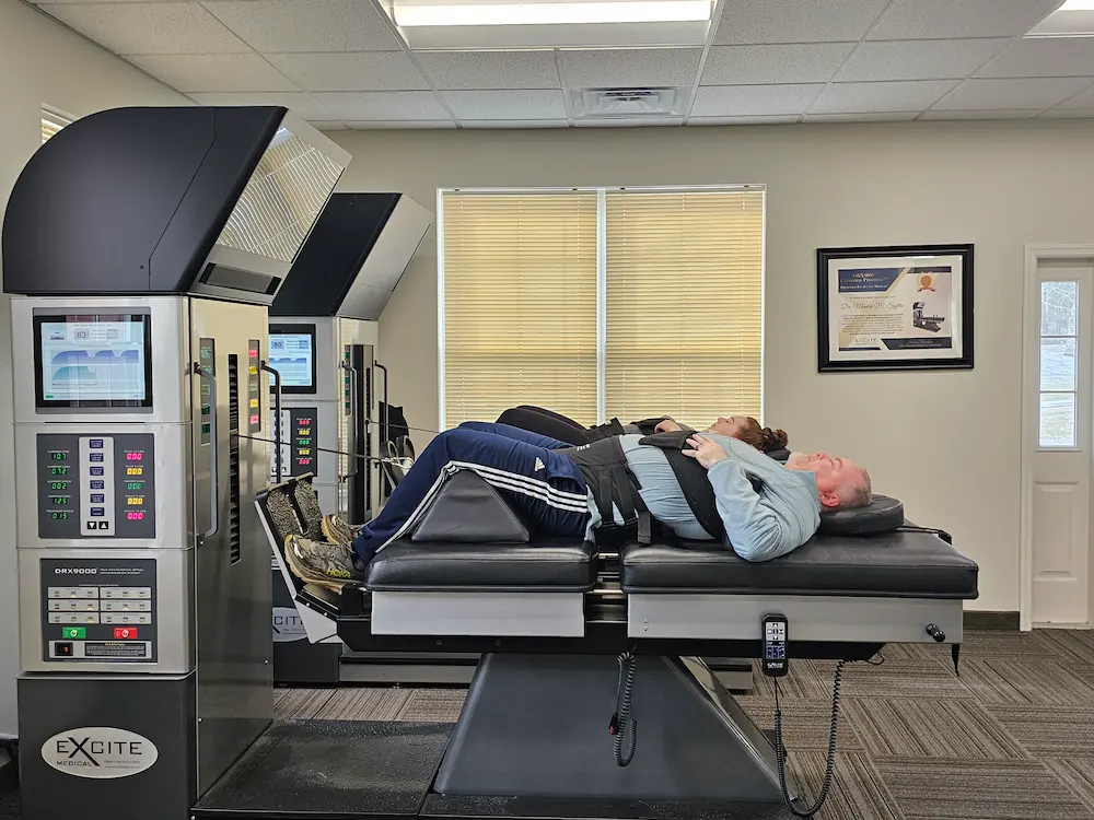 Two people lying on their backs on spinal decompression machines in a therapy room.
