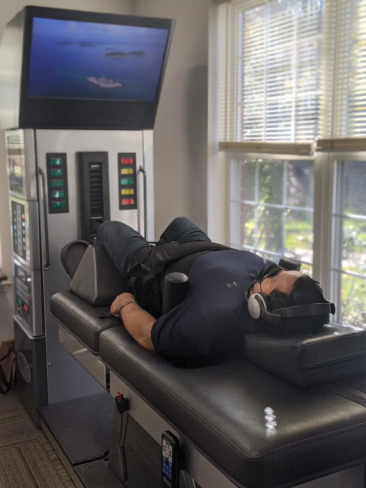 A man lies on a black therapy table with headphones, looking at a screen displaying an ocean scene, next to large windows.
