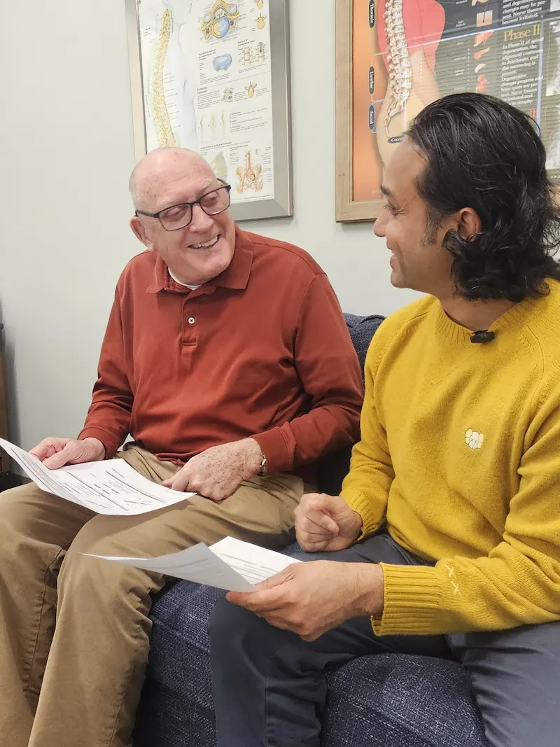 Two men, one older and one younger, are seated on a sofa, smiling and holding papers, with anatomical charts on the wall behind them.