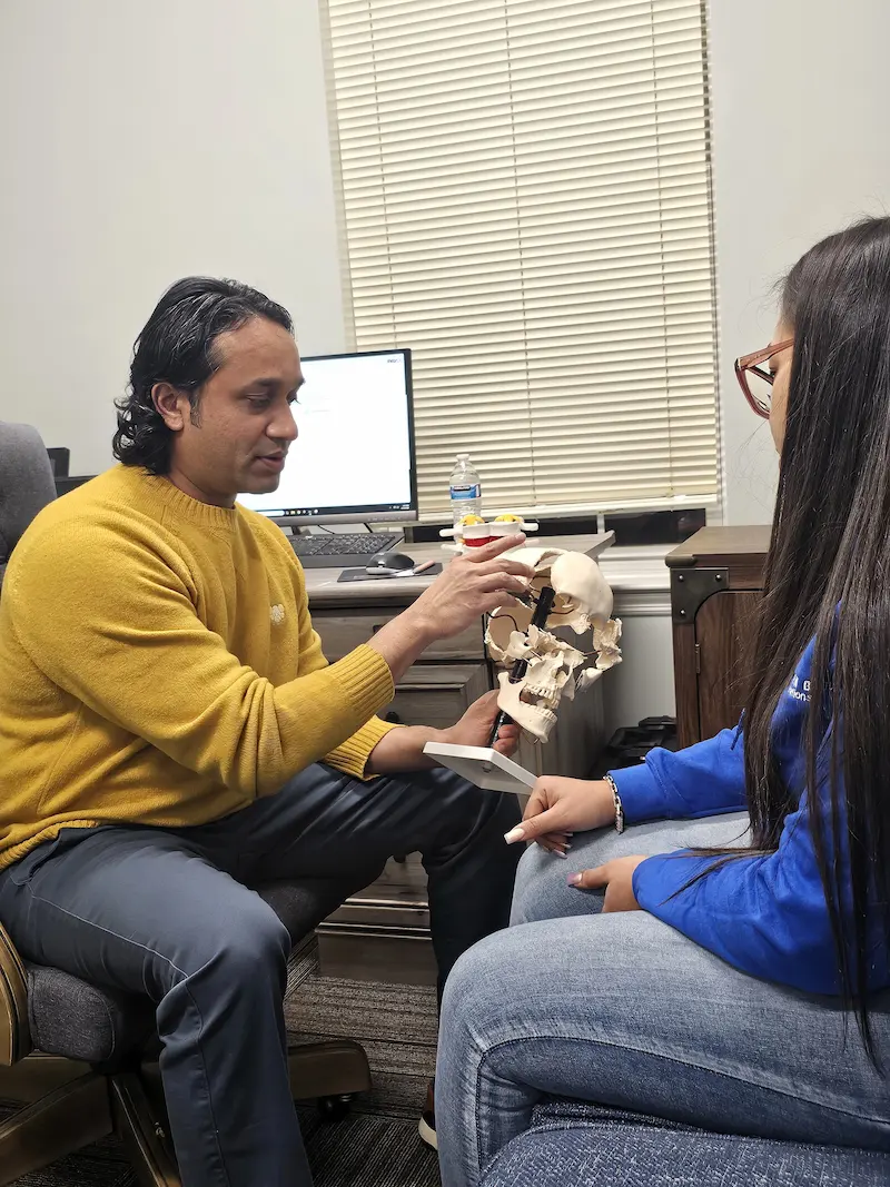 A man in a yellow sweater demonstrates a human skull model to a woman in a blue shirt, seated in an office.