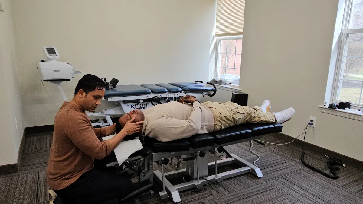 A chiropractor adjusts a patient's neck on a black treatment table in a room with windows.