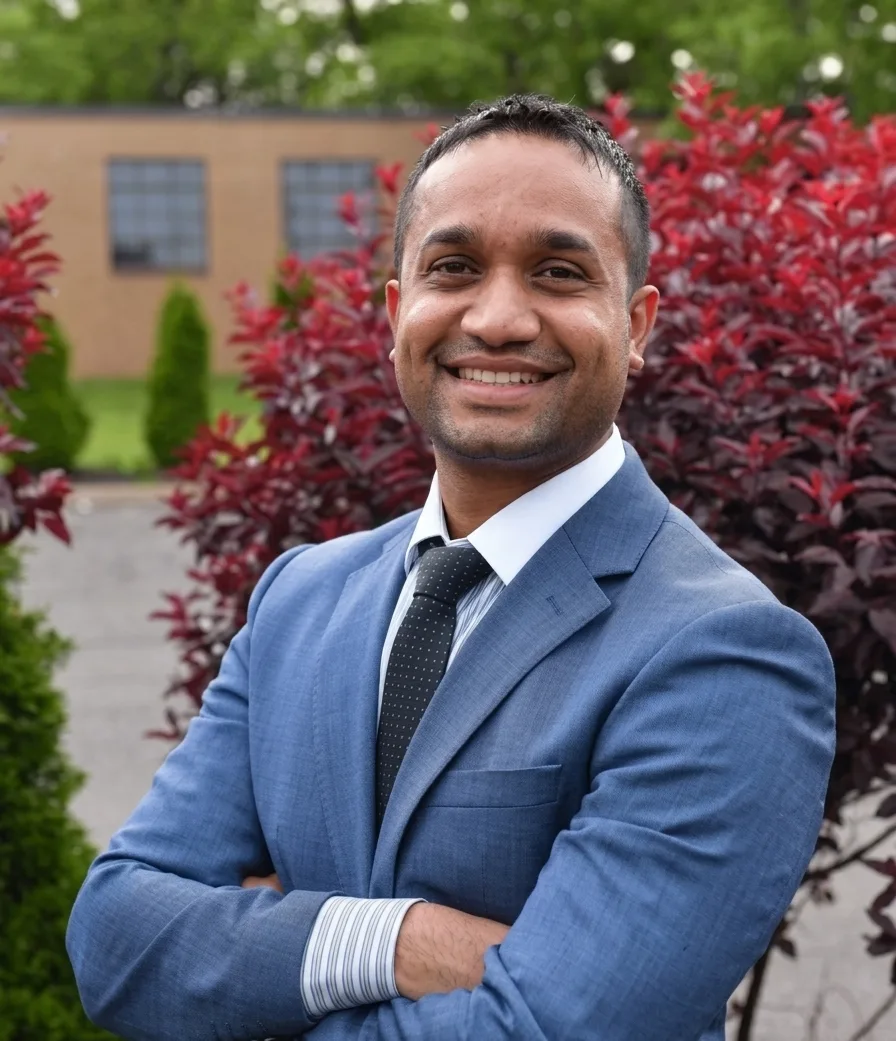 A man in a blue suit and tie, with arms crossed, smiling at the camera against a blurred background of red bushes and a building.