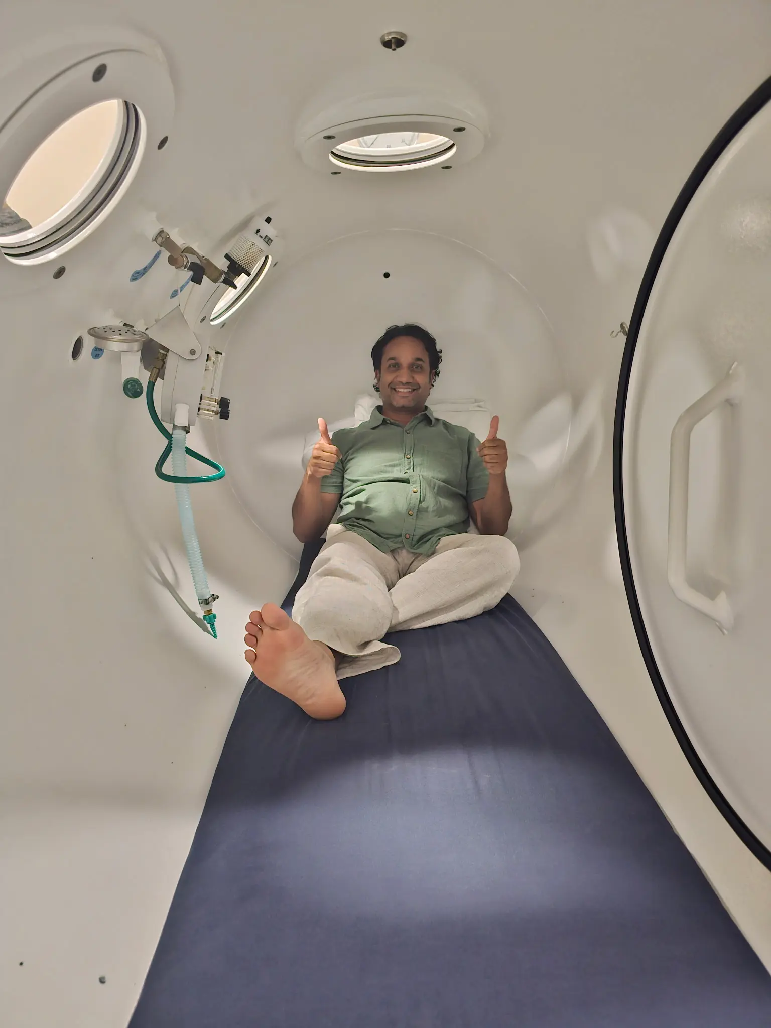 Man in a green shirt and light pants sitting cross-legged in a white hyperbaric chamber, smiling and giving two thumbs-up.