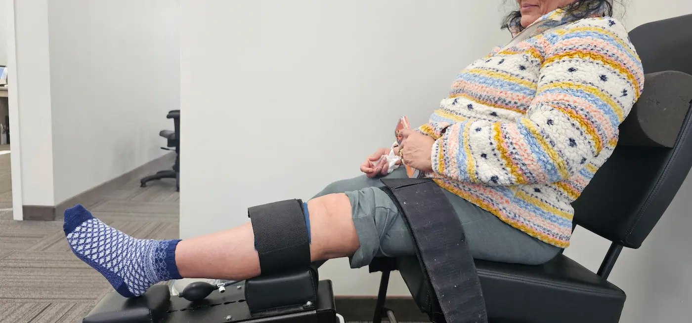 Side view of a person seated on a black therapy chair, with their left leg extended and secured to medical equipment. They wear a colorful jacket and blue patterned socks.