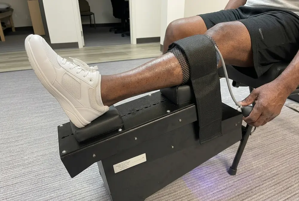 A person's right leg resting on a black medical device, with a strap around the calf and a hand pump being operated.