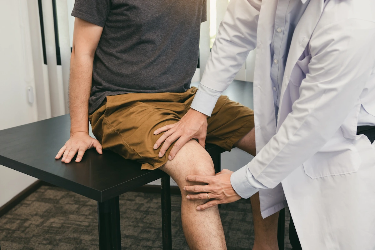 A doctor in a white lab coat examines a patient's right knee with both hands. The patient is seated on a black table, wearing a grey t-shirt and brown shorts.