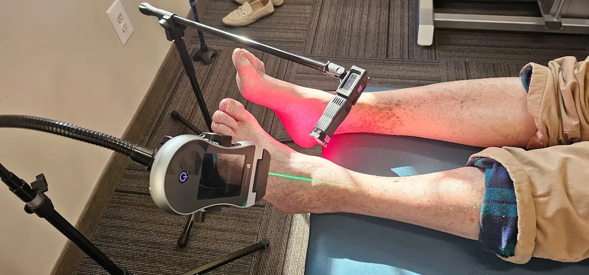 A person's legs and feet undergoing laser therapy. One device emits a red light on the foot, and another emits a green laser line on the shin.