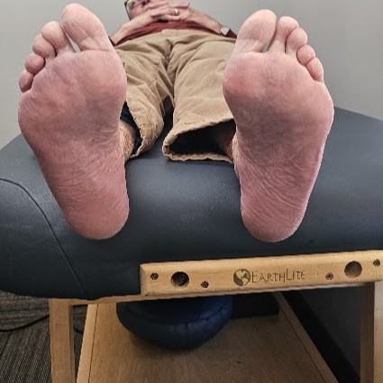 A person's bare feet, soles facing up, resting on a dark blue examination table with a wooden frame.