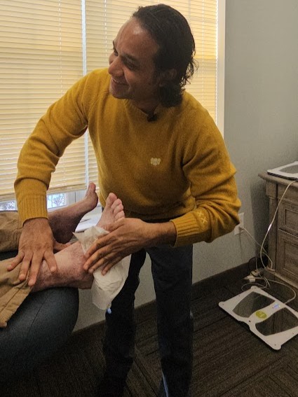 A smiling man in a yellow sweater attends to a person's lower leg and foot with a white cloth.