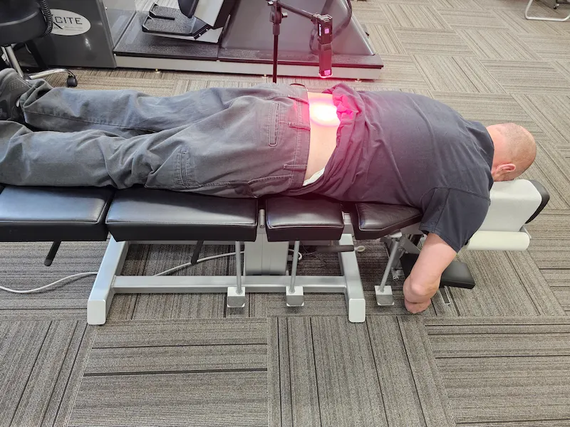 A man lies face down on a black treatment table, receiving red light therapy on his lower back from an overhead device.