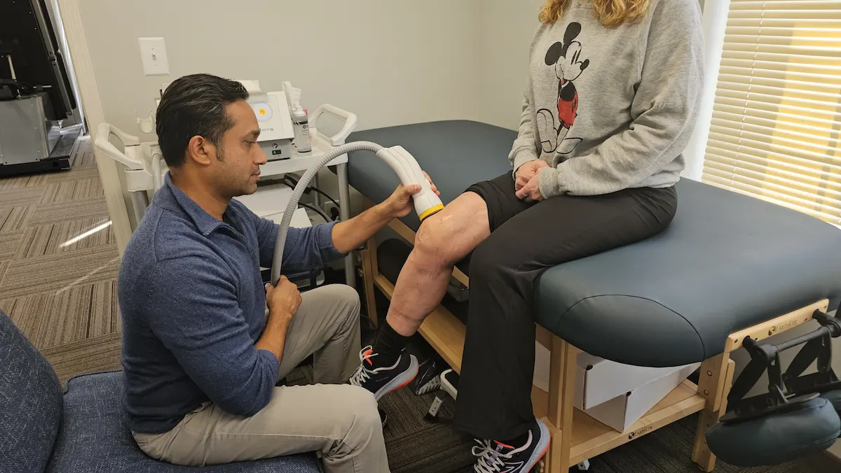 A man applies a medical device to a woman's knee as she sits on an examination table.