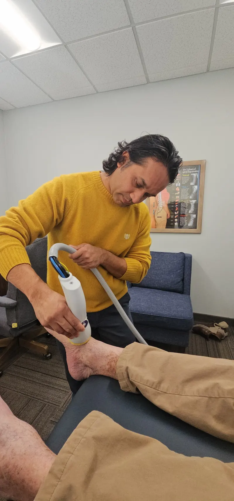 A male therapist uses a handheld device to treat a patient's ankle in a clinical setting.