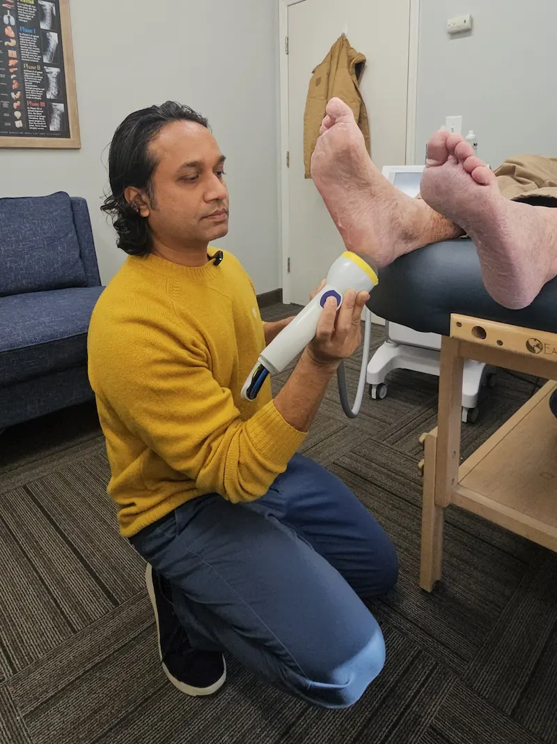 A man kneels, applying a white and yellow handheld device to the sole of a patient's foot on an examination table.