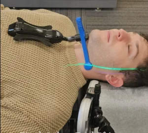 A man lies on his back with a blue jaw support and a device on his chest, showing green and purple laser lines across his neck.