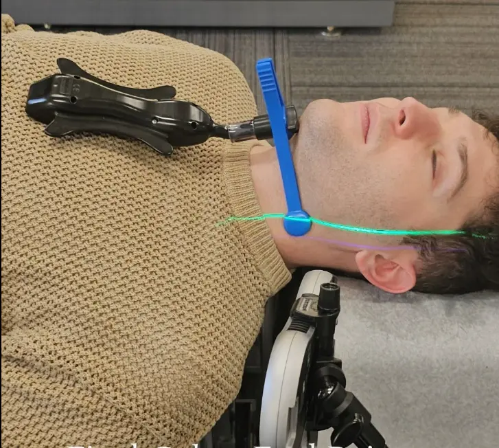 A man lies on his back with a blue jaw support and a device on his chest, showing green and purple laser lines across his neck.
