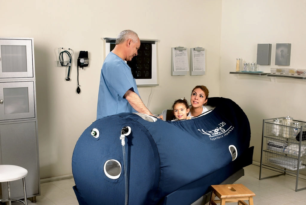 A man in scrubs attends to a woman and child inside a blue hyperbaric chamber in a medical room.