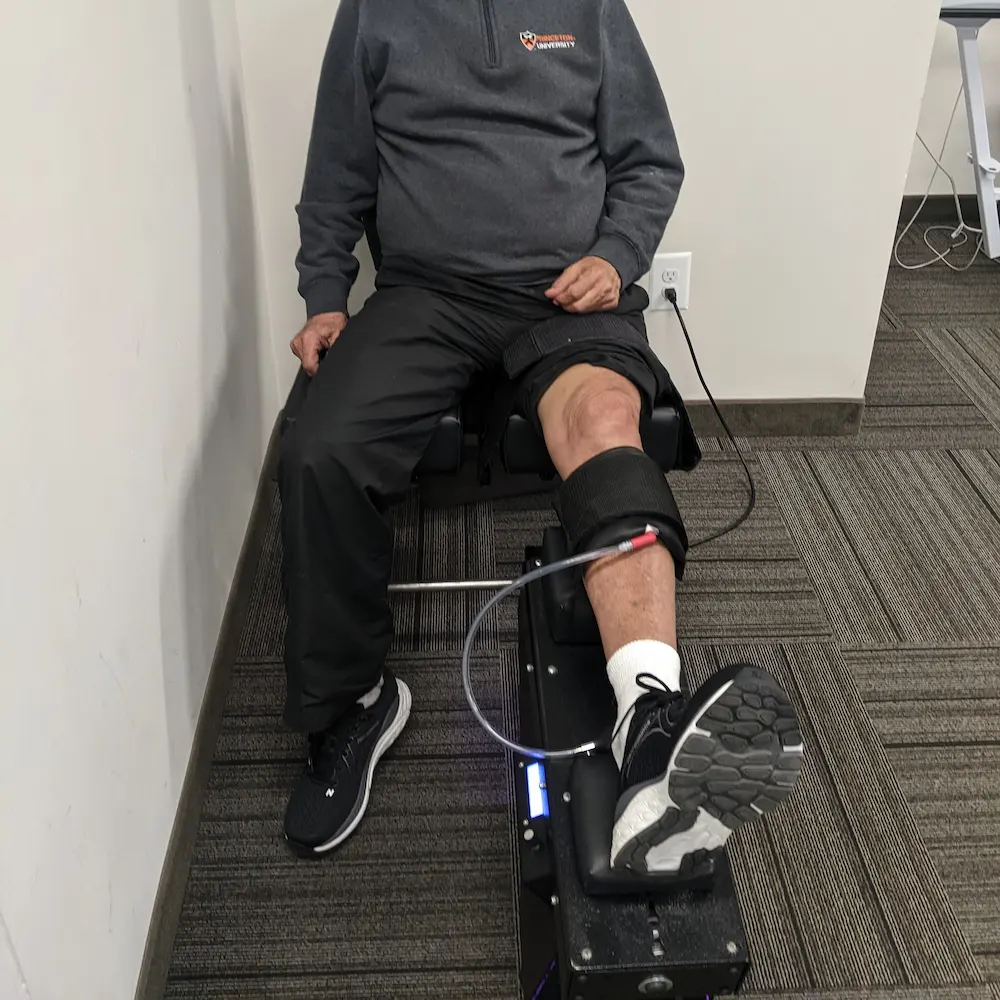 A person seated, with their right leg strapped into a rehabilitation machine, wearing a Princeton University sweatshirt.