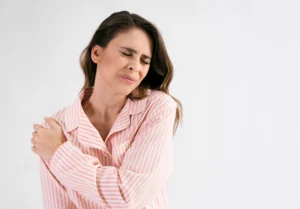 A woman in pink striped pajamas holds her shoulder with a pained expression.