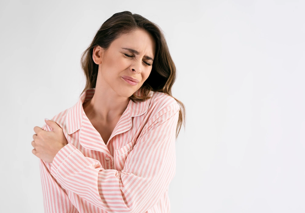 A woman in pink striped pajamas holds her shoulder with a pained expression.