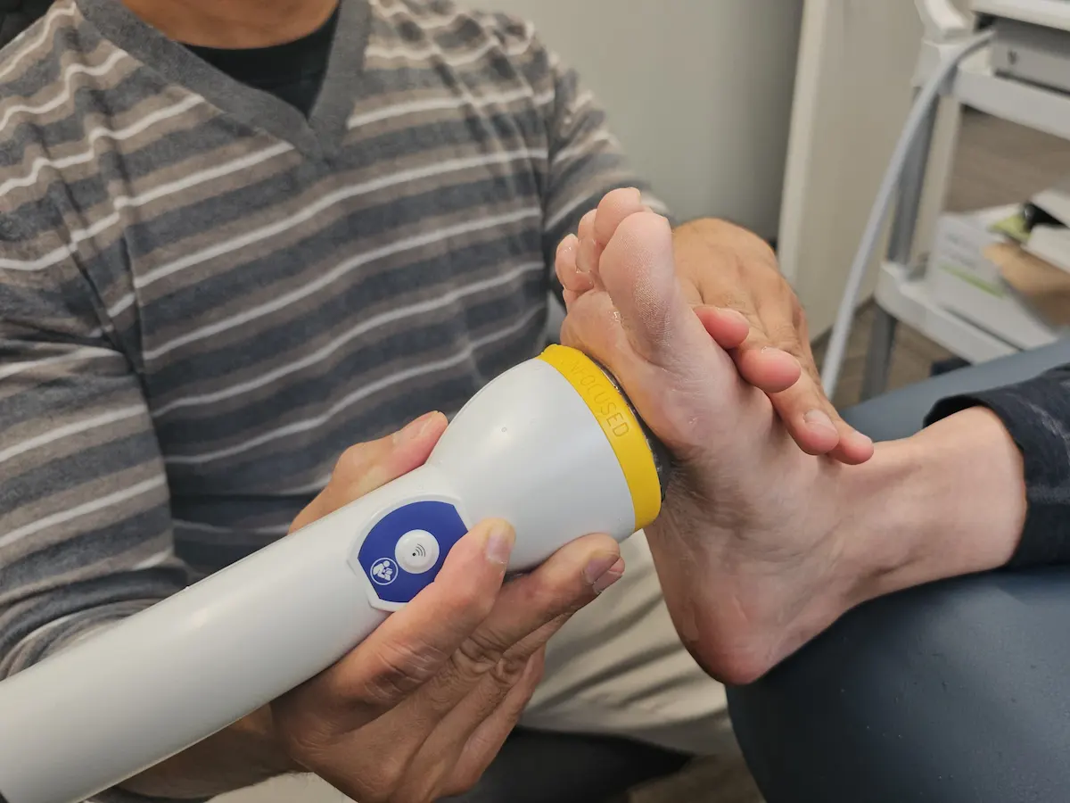 A person's hands hold a white and yellow handheld device against the sole of another person's bare foot.